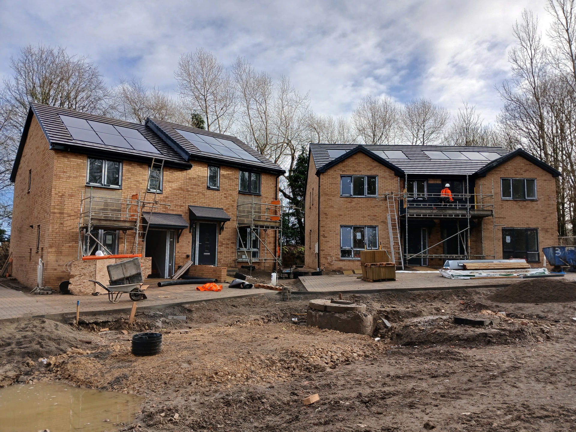 Four newly constructed semi-detached brick houses on a muddy building site. The houses have solar panels on the roof and windows covered in protective film. The buildings have scaffolding surrounding them and a person in a high-visibility jacket and hard hat is stood on the second level on one of the scaffolding structures facing toward the building. Construction materials and equipment are scattered around the site.