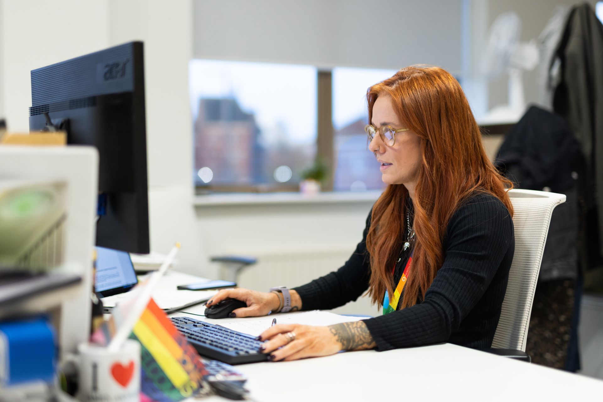 A woman with long red hair and glasses working at a desk in an office, focused on her computer screen. She is wearing a black top, a rainbow lanyard, and has tattoos on her arms. On the desk are a keyboard, mouse, and a mug with a rainbow design that says 'I ❤️'. A rainbow pride flag is also visible, along with other office supplies. A window in the background shows an urban cityscape