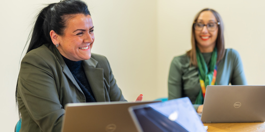 Two people sat next to other other at a table with laptops. The person closest to the camera is in focus and is smiling joyfully.