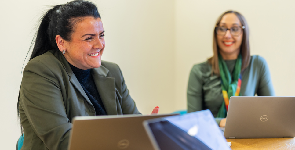 Two people sat next to other other at a table with laptops. The person closest to the camera is in focus and is smiling joyfully.