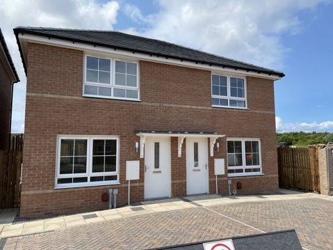 A modern red-brick semi-detached house with two front doors, white framed windows, and a shared paved driveway in front. Each home has a small canopy above the entrance and fenced gardens to the side.