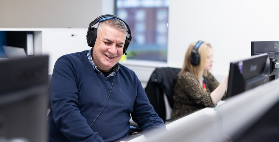 A man sat at a desk in front of a computer smiling with a headset on. 