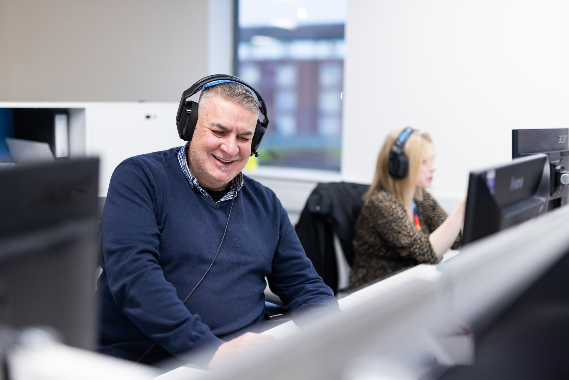 A man sat at a desk in front of a computer smiling with a headset on. 