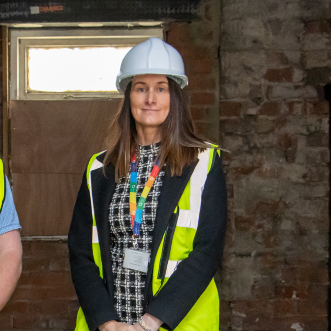 A woman smiling proudly wearing a hard hard and high vis vest. She is standing against an exposed brick wall.