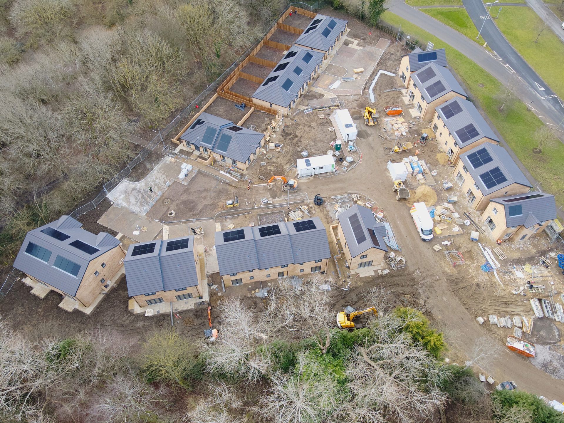 Aerial view of a housing development under construction, surrounded by woodland and a nearby road. The site features several newly built brick homes with solar panels on the roofs, construction vehicles, temporary site cabins, and building materials scattered around. The layout forms a small, planned community with pathways and shared spaces taking shape.