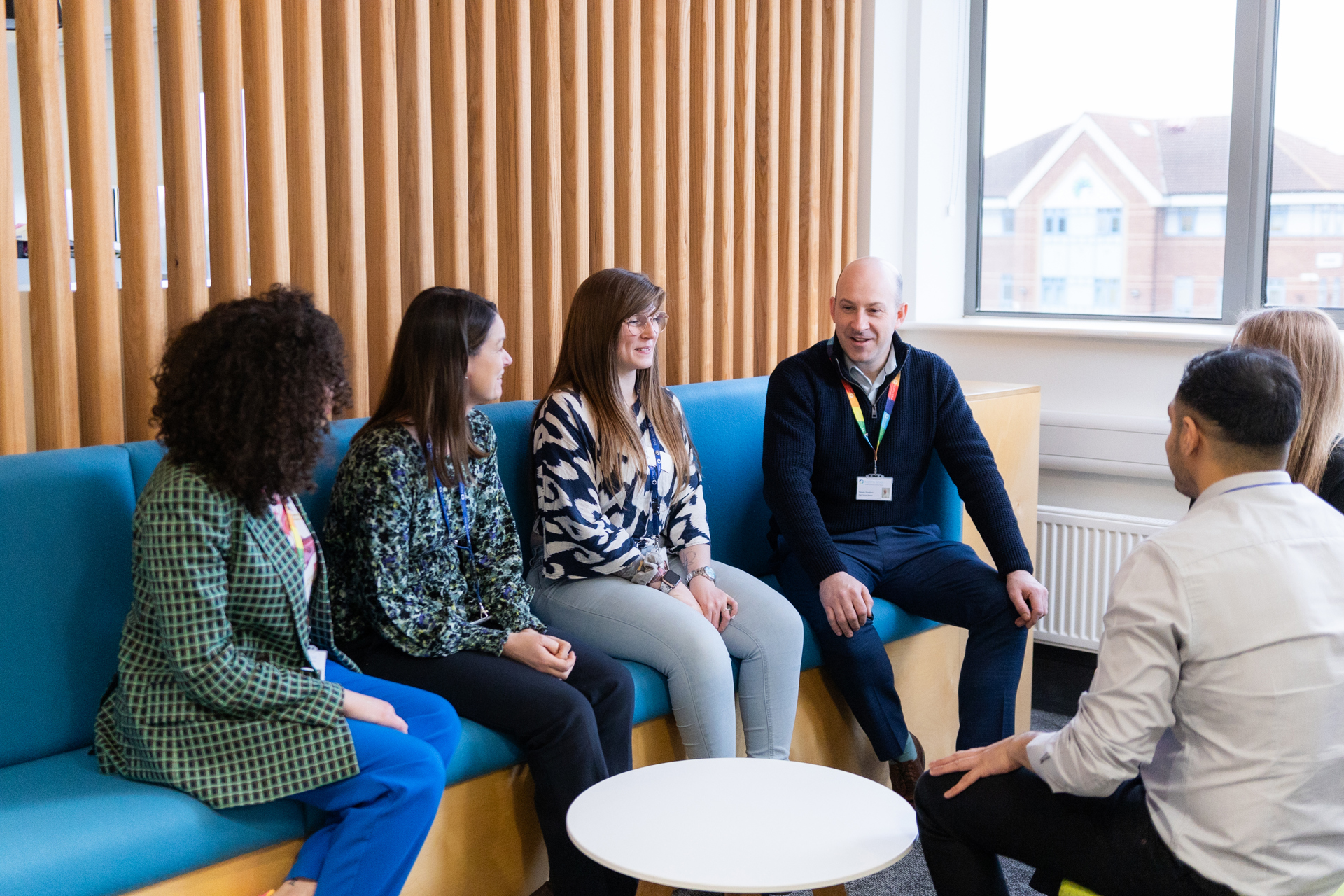 A group of six people sit in a casual meeting space at North Star's office. They are seated on blue cushioned benches around a small white table, engaged in conversation. A wooden slat wall and large window with a view of nearby buildings create a bright, welcoming atmosphere. Everyone appears relaxed and focused.