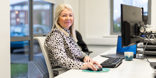 A woman with long blonde hair sits at a desk in an office, smiling at the camera. She is wearing a patterned black and white blouse and using a computer mouse. A keyboard, monitor, and a blue and white mug are on the desk in front of her. Large windows in the background show a car park and buildings outside